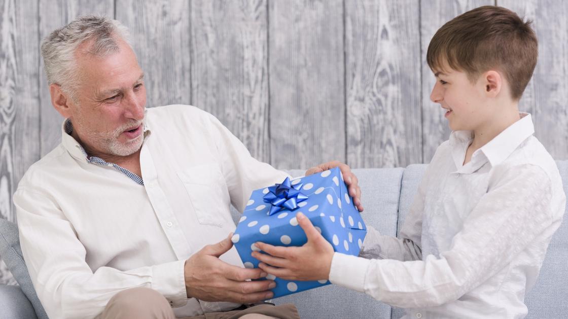 Мальчик вручает седому мужчине подарочную коробку в синей упаковке с белым горошком