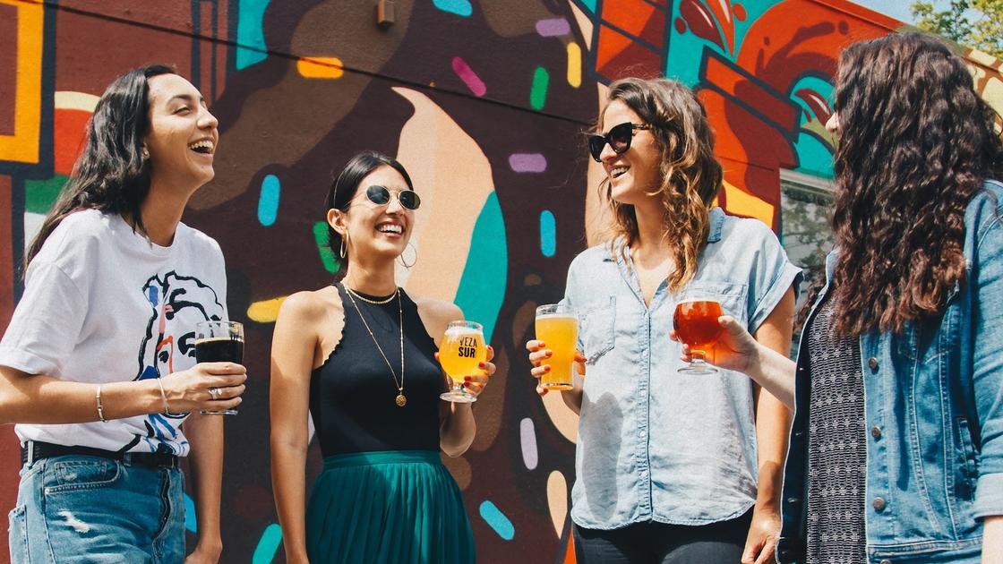 Четыре девушки с бокалами в руках стоят возле разрисованной стены. Смеющиеся девушки одеты по-летнему в практичные наряды, две из них в солнцезащитных очках