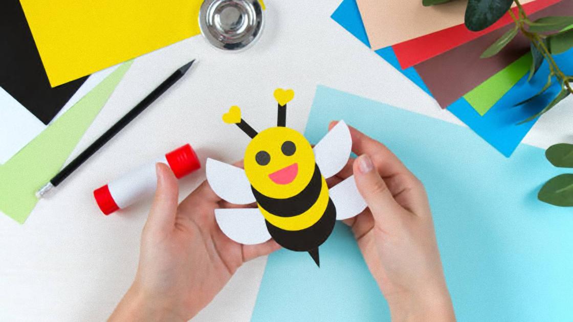 В руках над столом держат пчелку, сделанную из бумаги. На столе лежат листы цветной бумаги, карандаш, клей