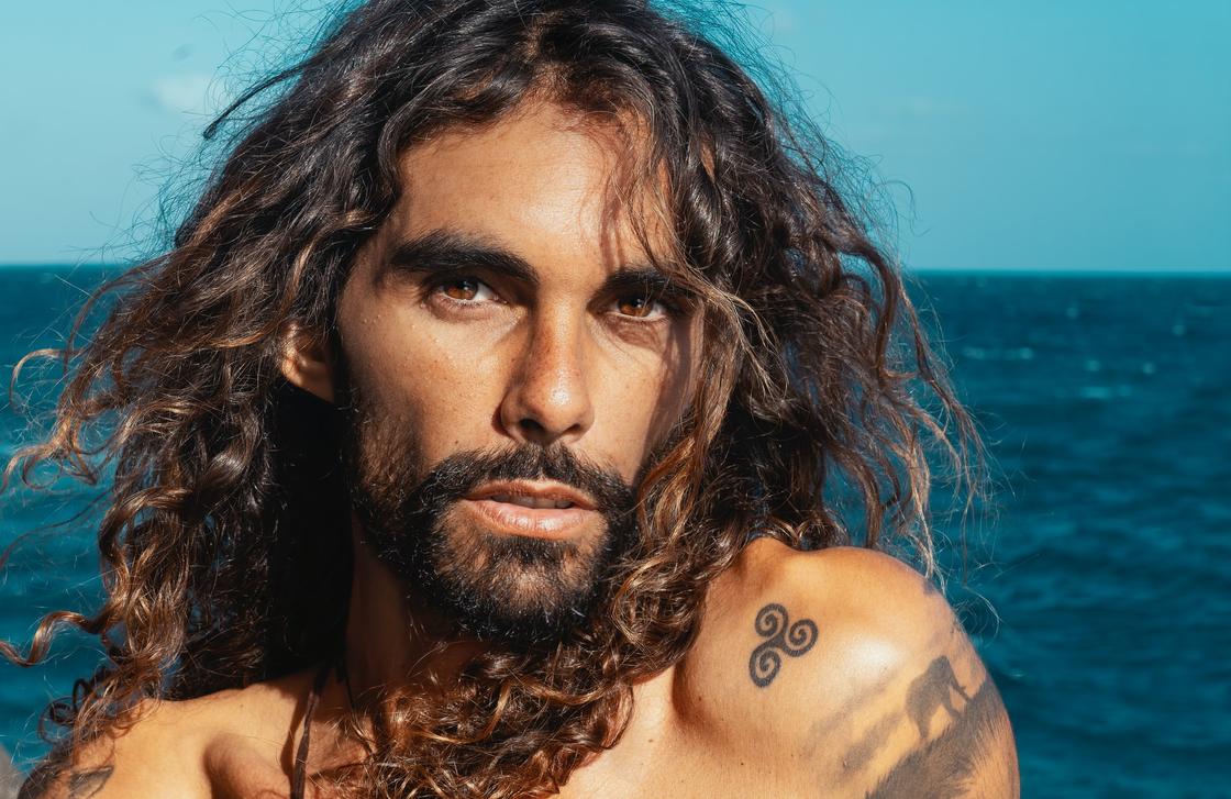 Мужчина с длинными вьющимися волосами и бородой смотрит в камеру на фоне моря