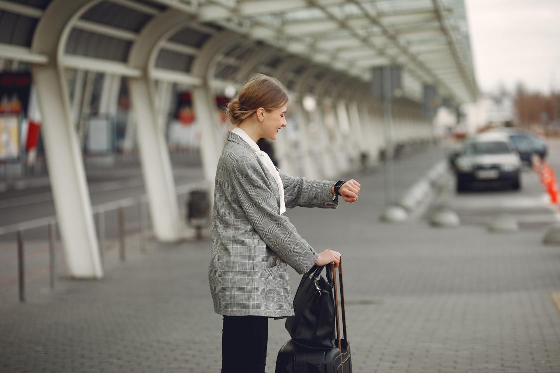 Девушка стоит с чемоданом у зоны парковки аэропорта и смотрит на часы на руке