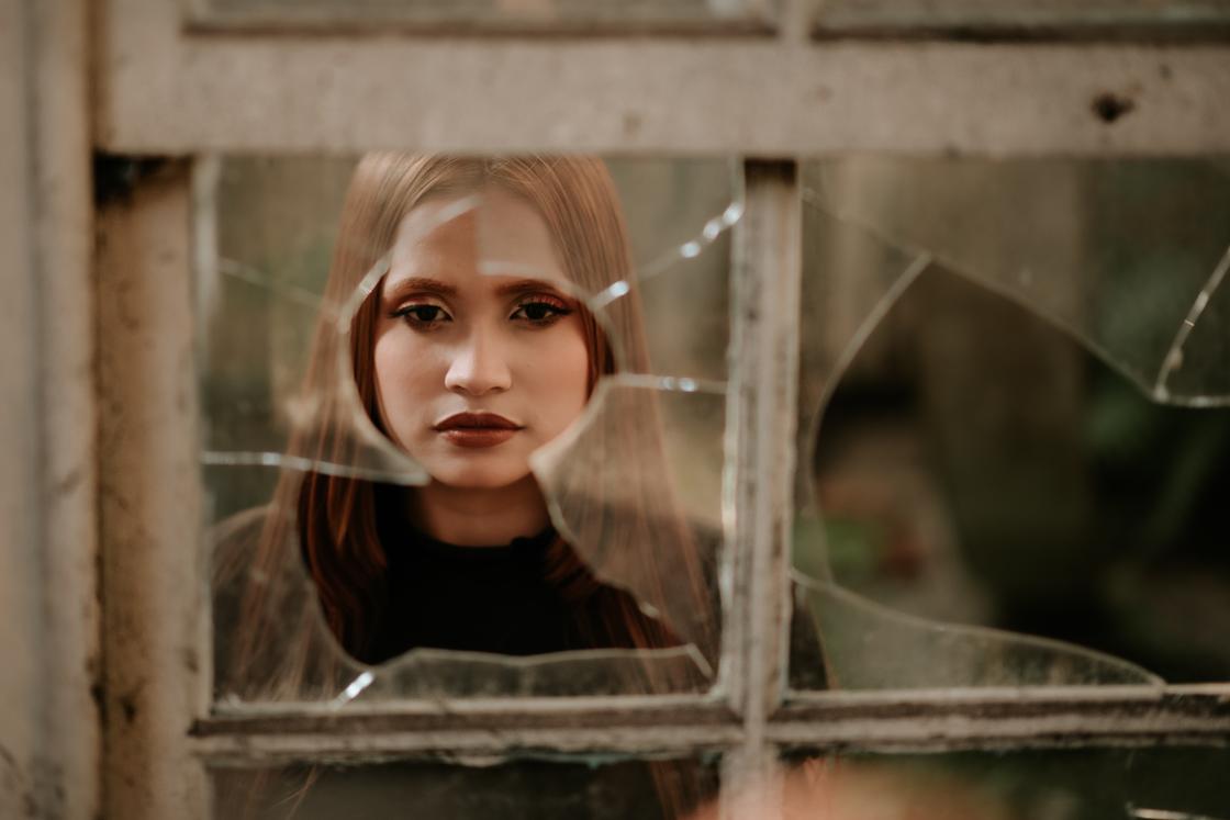 Девушка смотрит в камеру через дыру в разбитом окне