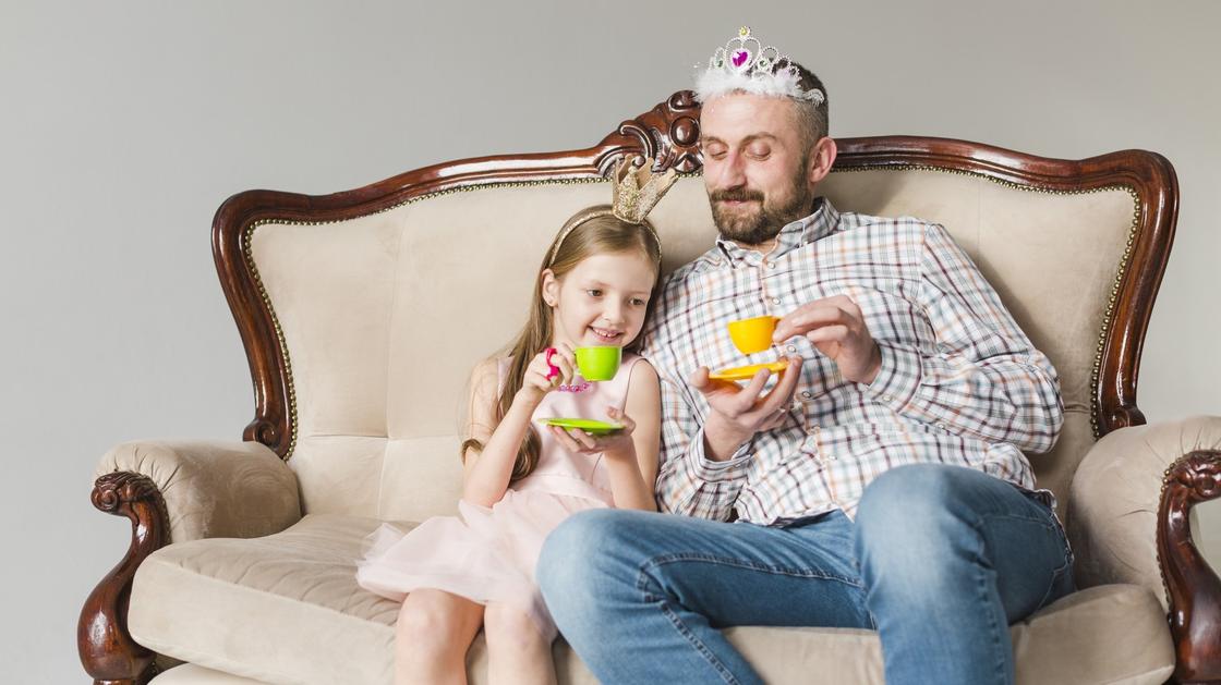 Папа с дочкой в коронах сидят на диване и пьют с игрушечных чашек