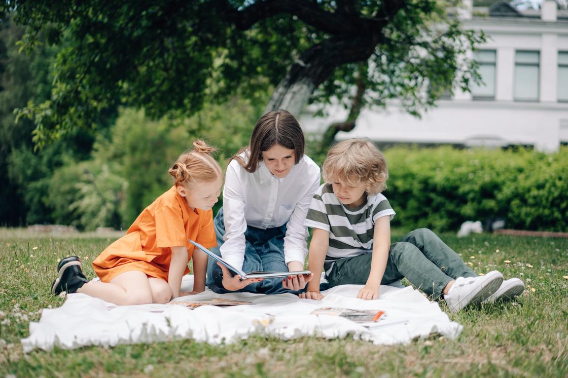 Мама с детьми читает книгу, сидя на покрывале на траве