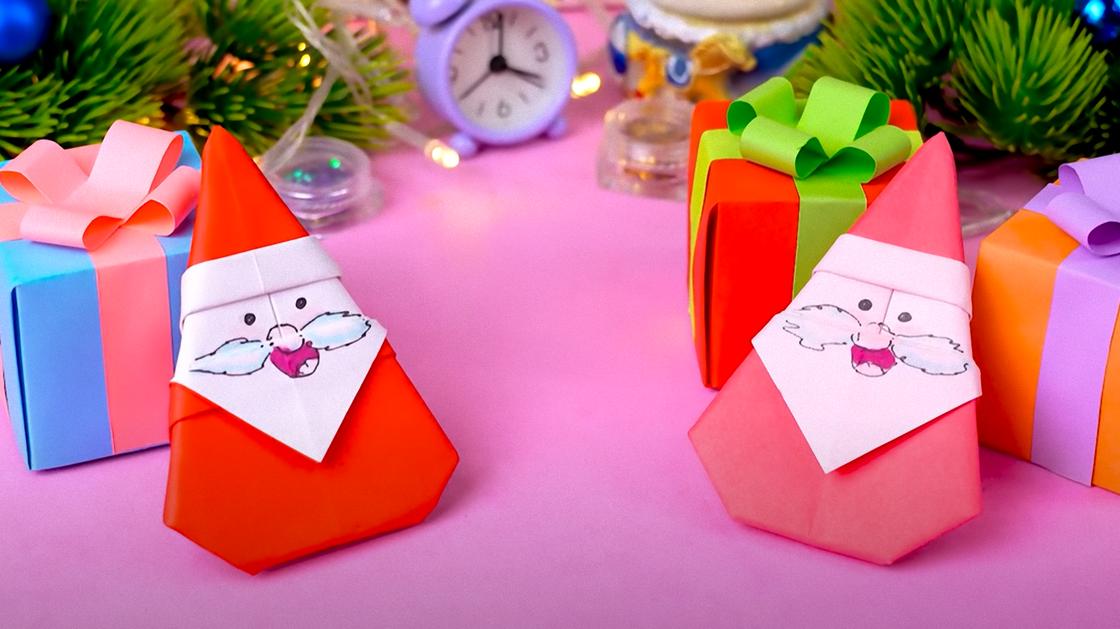 Возле новогодней елки на столе лежат цветные коробочки и две фигурки Деда Мороза, сделанные по технике оригами