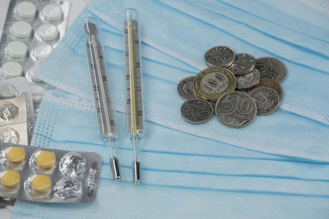 Монеты тенге лежат на медицинских масках рядом с градусниками