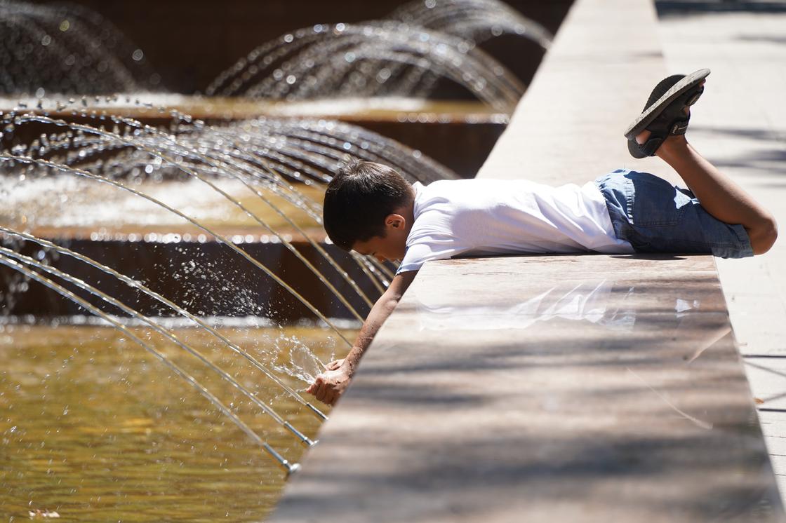 Мальчик лежит на бортике фонтана и трогает воду руками