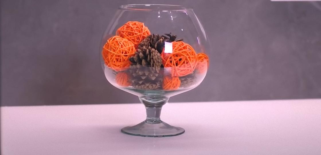 В стеклянной вазе на высокой ножке размещены сосновые шишки и оранжевые шарики из полосок бумаги