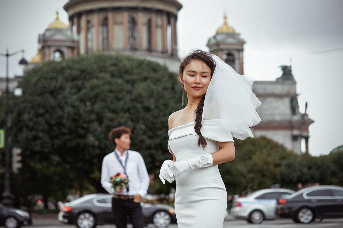 Невеста в белом платье на фоне жениха, машин и городского пейзажа