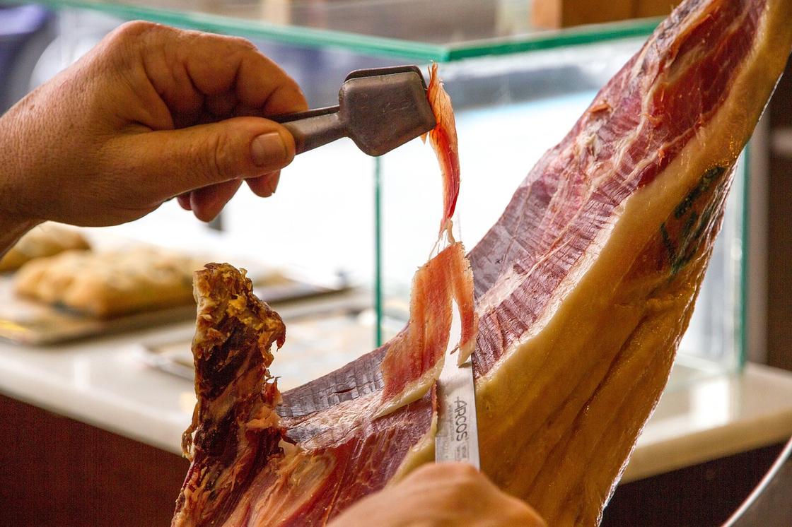 Мужчина нарезает свиной окорок тонким ножом и при помощи щипцов держит кусочек в руках