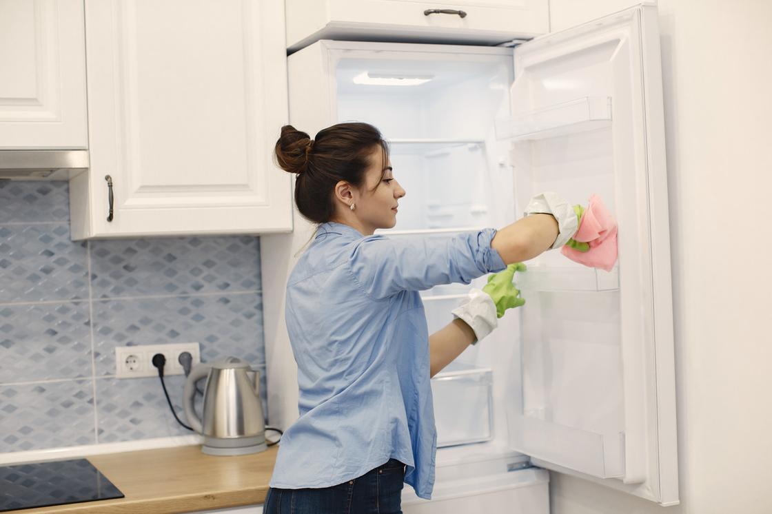 Девушка в резиновых перчатках вытирает розовой салфеткой внутреннюю часть дверцы холодильника