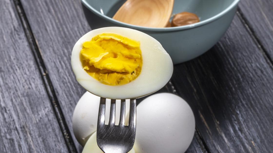 На вилке надета половинка вареного яйца. На столе лежат еще яйца в скорлупе и стоит миска с деревянной лопаткой