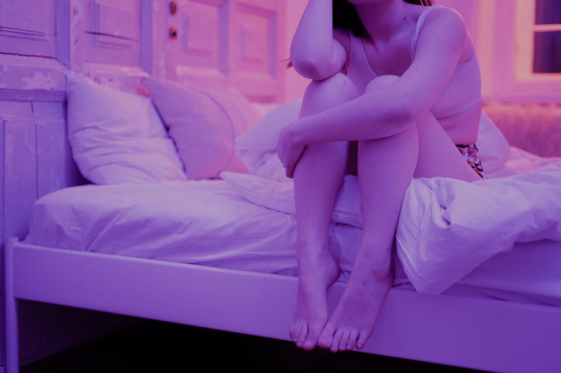 Полураздетая девушка сидит в кровати при свете фиолетовой лампы