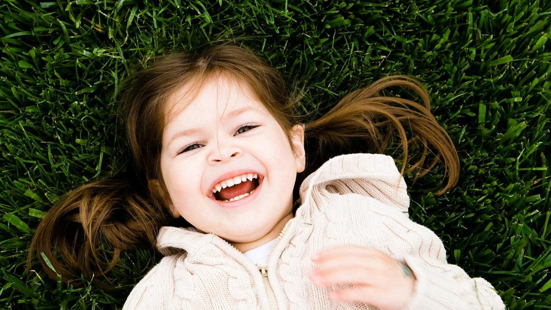 Девочка с хвостиками в теплом вязаном свитере лежит на зеленой траве и широко улыбается
