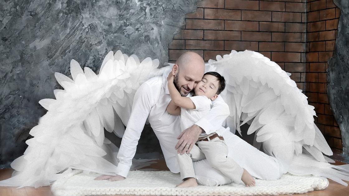 Мальчик обнимает папу. Они сидят на коврике, а за ними лежат большие белые крылья
