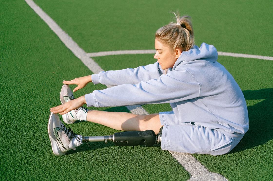 Девушка с протезом на левой ноге занимается растяжкой, сидя на футбольном поле