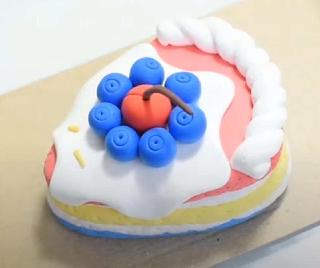 Кусок слоеного торта из воздушного пластилина украшен синими ягодами, красной ягодой и белым кремом