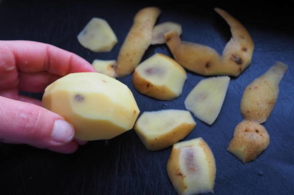 Очищенный картофель с темными пятнами держат в руке. На столе лежат обрезки картофеля с пятнами