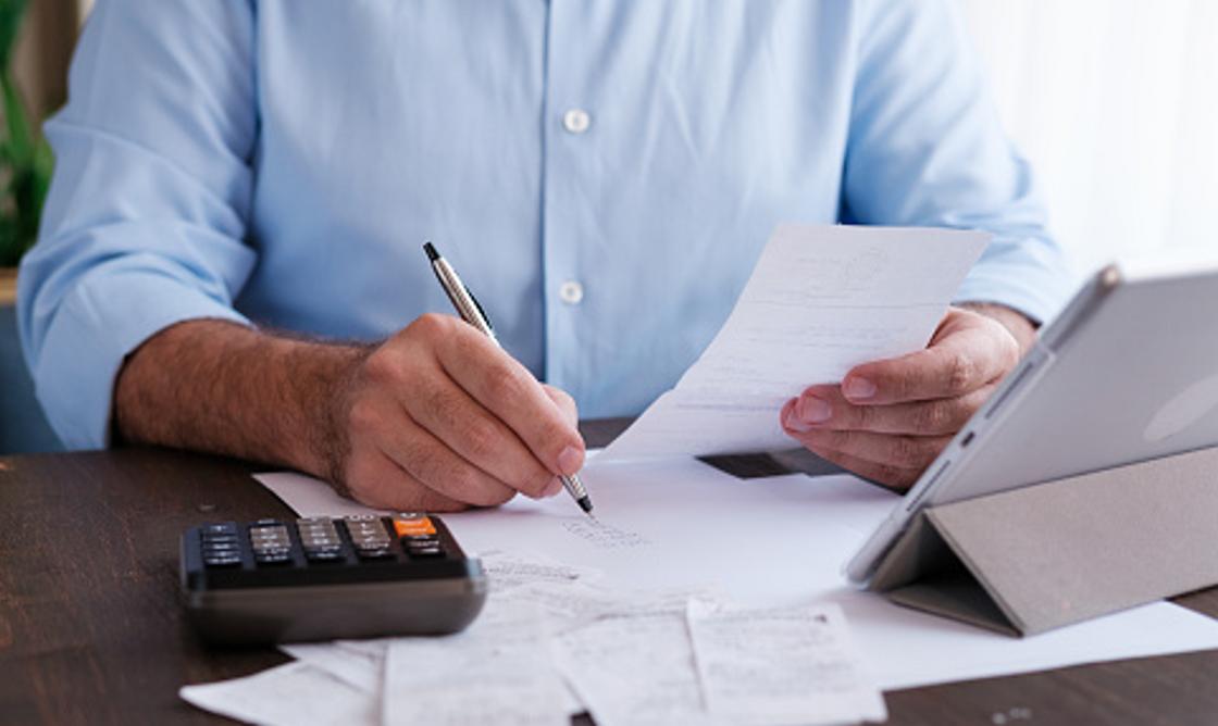 Мужчина держит счет, ручку, перед ним чеки, калькулятор и планшет
