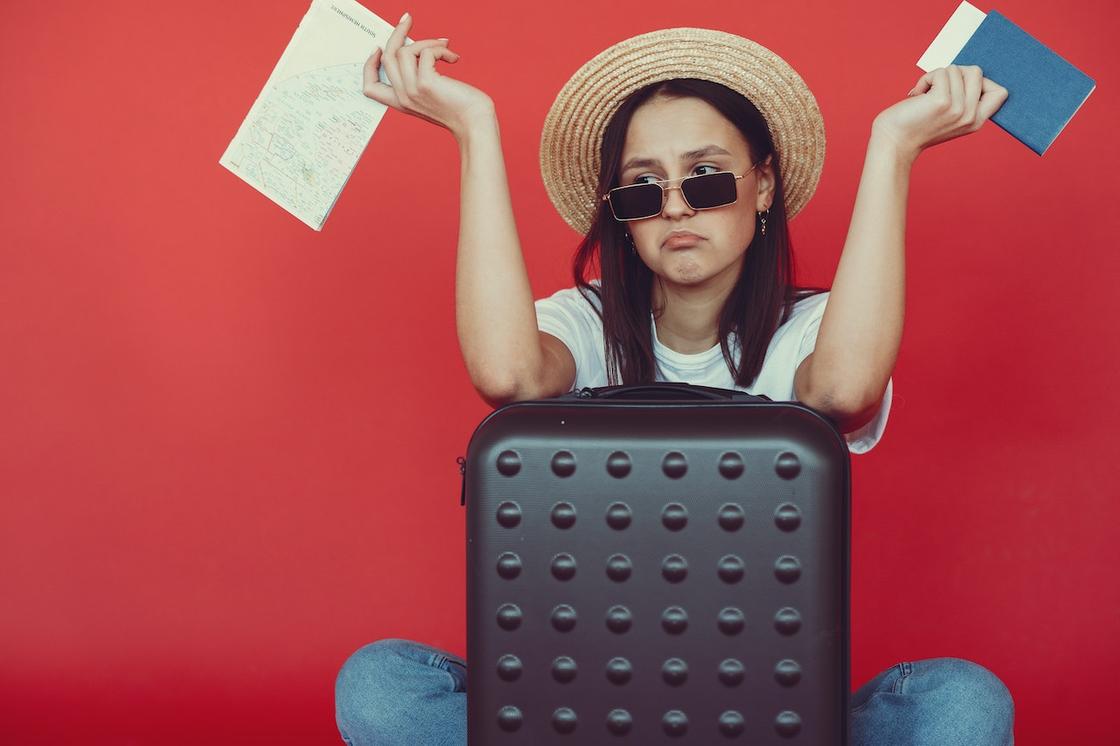 Огорченная девушка в шляпе с билетами, паспортом и чемоданом на красном фоне
