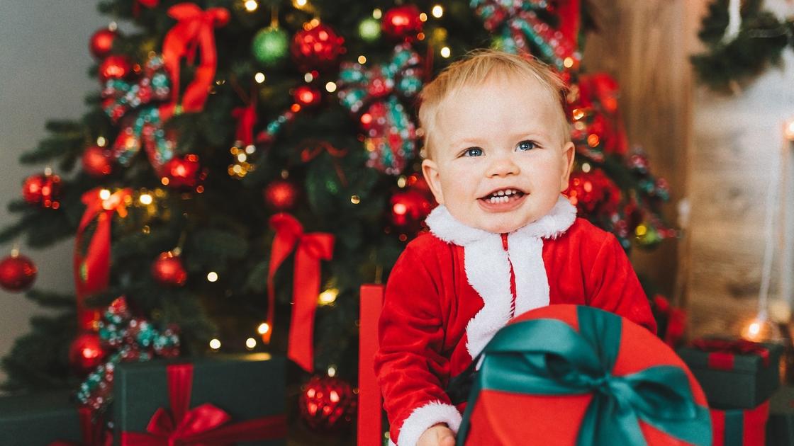 Под наряженной елкой сидит улыбающийся мальчик в костюме Санта-Клауса и держит в руках круглую красную коробку, перевязанную зеленой лентой