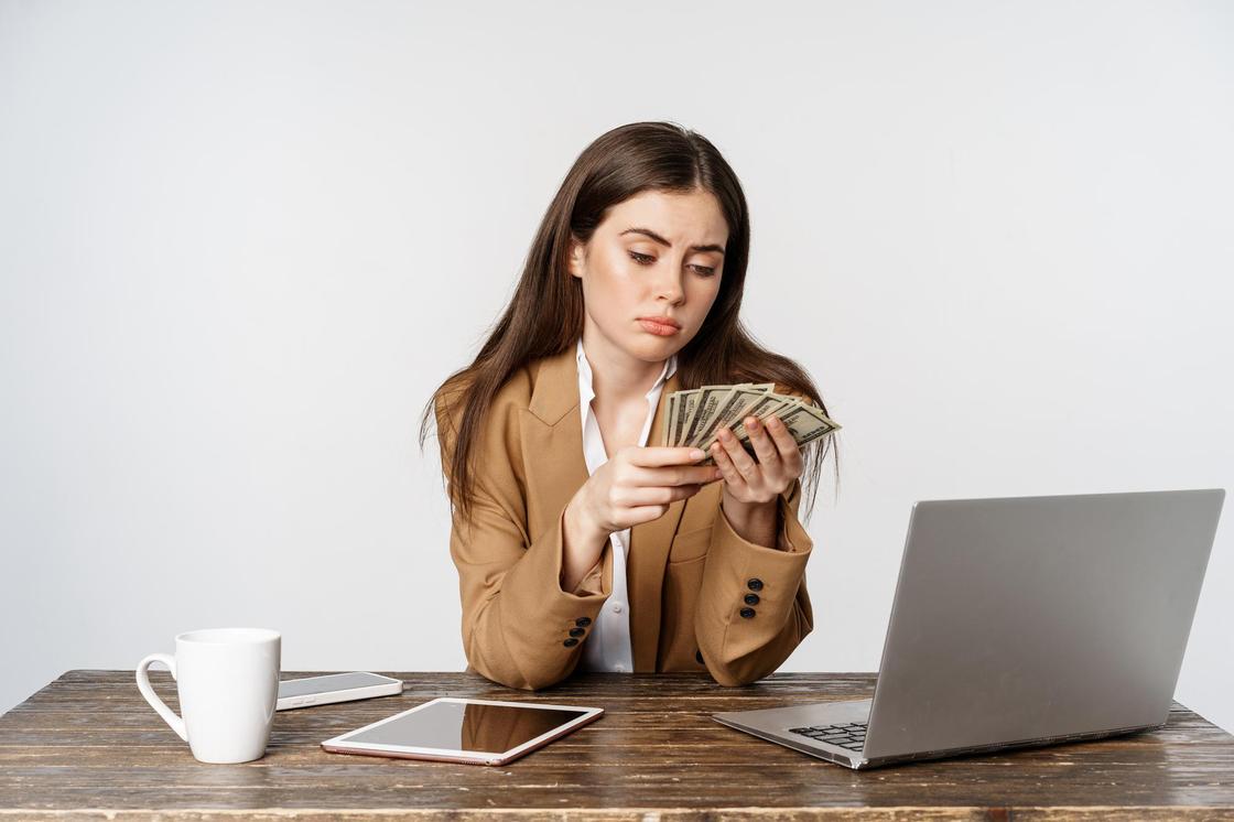 Девушка с озадаченным лицом сидит перед ноутбуком и держит в руках доллары