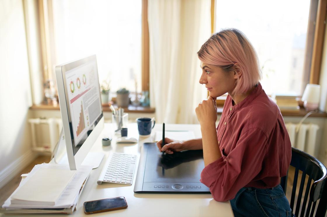 Девушка с розовыми волосами сидит перед монитором и держит стилус на графическом планшете