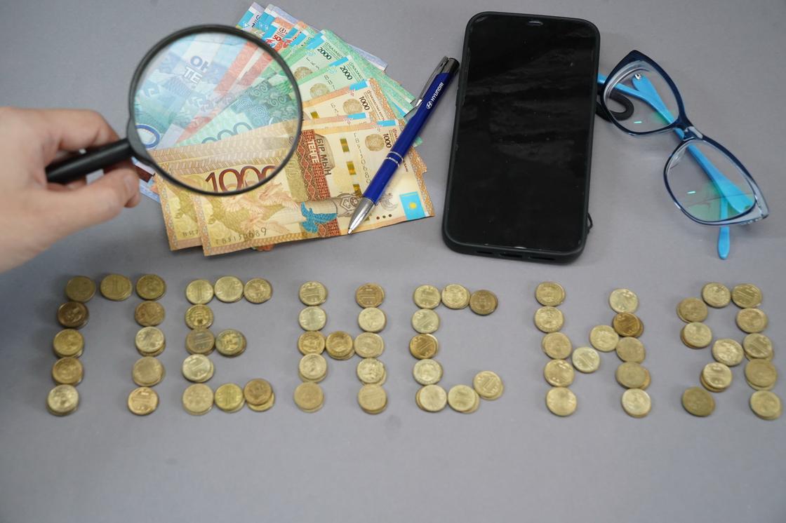Монеты и купюры тенге лежат рядом со смартфоном и очками