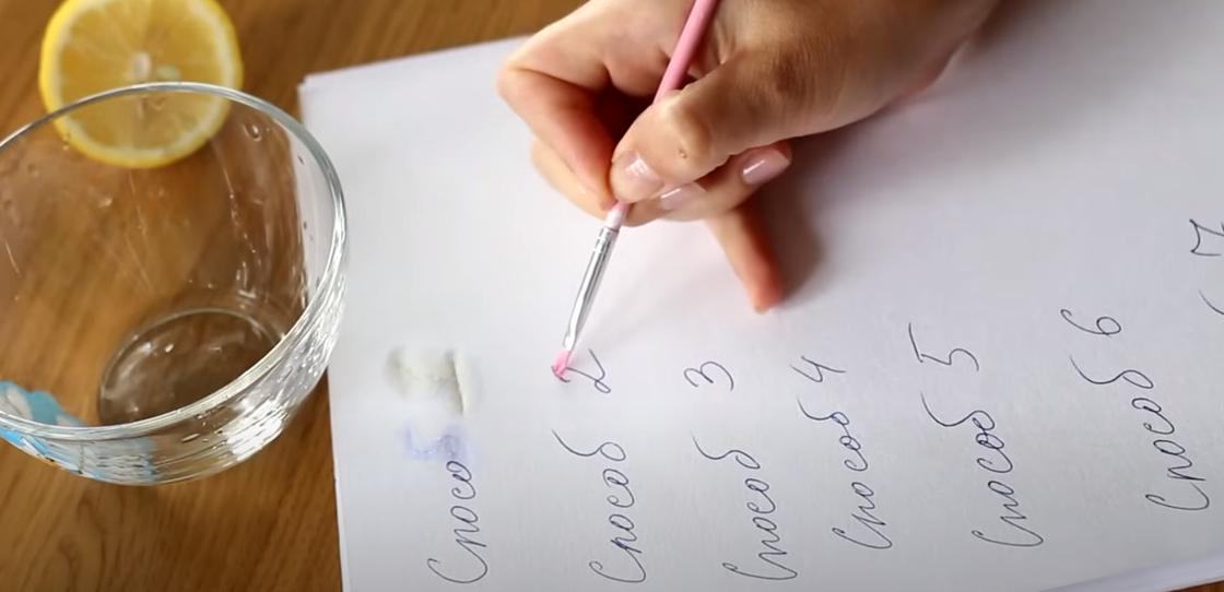 На белом листе бумаги кисточкой проводят по надписям, сделанным ручкой. Рядом стоит стеклянная пиала и лежит половина лимона