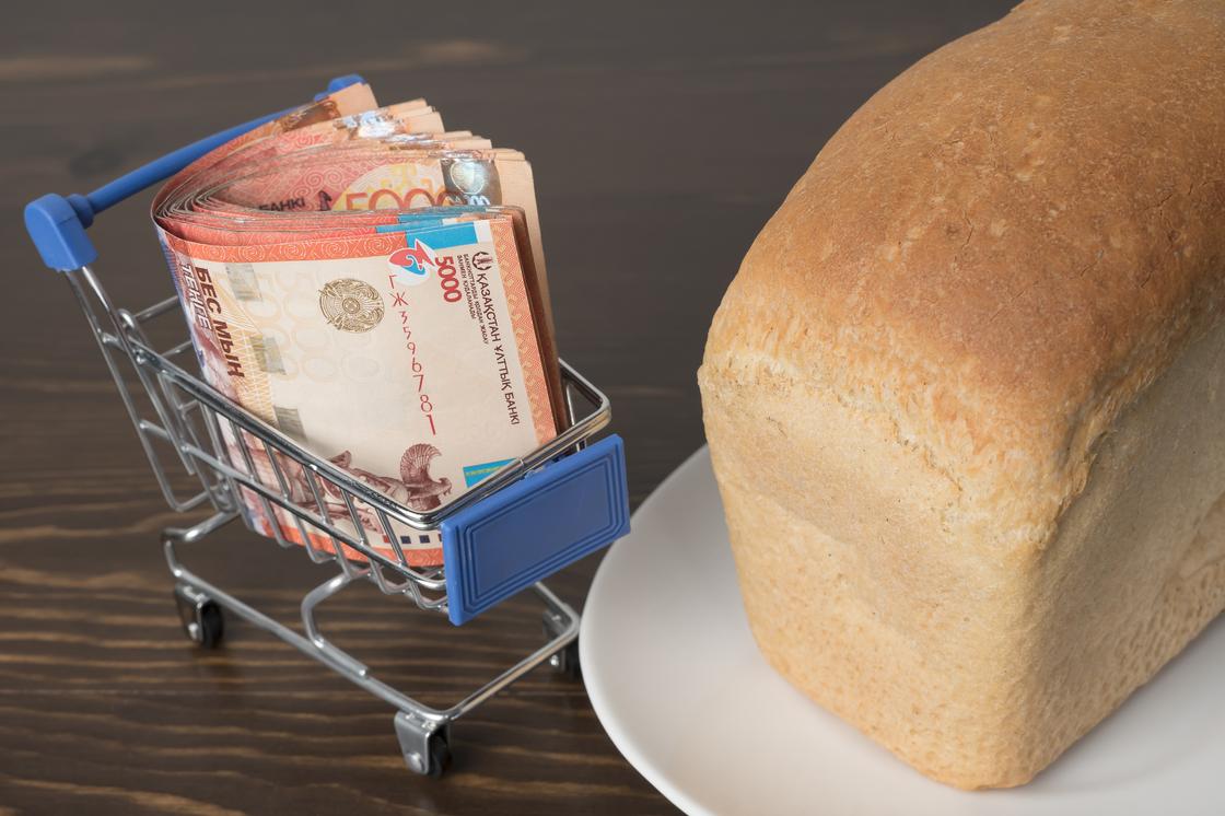 Пачка тенге лежит в миниатюрной корзине рядом с булкой хлеба