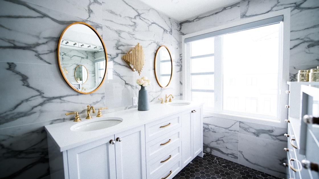 Ванная комната с яркой позолоченной фурнитурой, двойной белой тумбой с раковинами, круглыми зеркалами и оригинальным светильником. Стены обложены мраморной плиткой