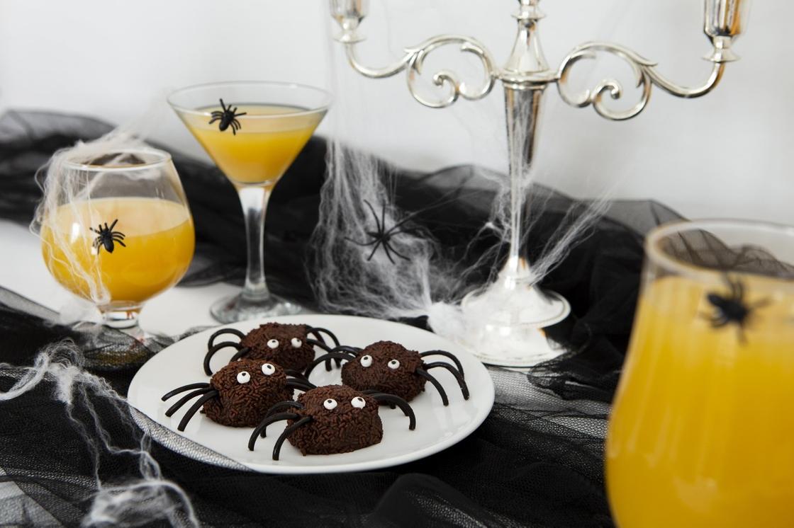 На столе стоит белая тарелка с четырьмя пирожными-пауками, подсвечники и бокалы с желтым напитком. Все приборы обтянуты паутиной и на них сидят черные паучки