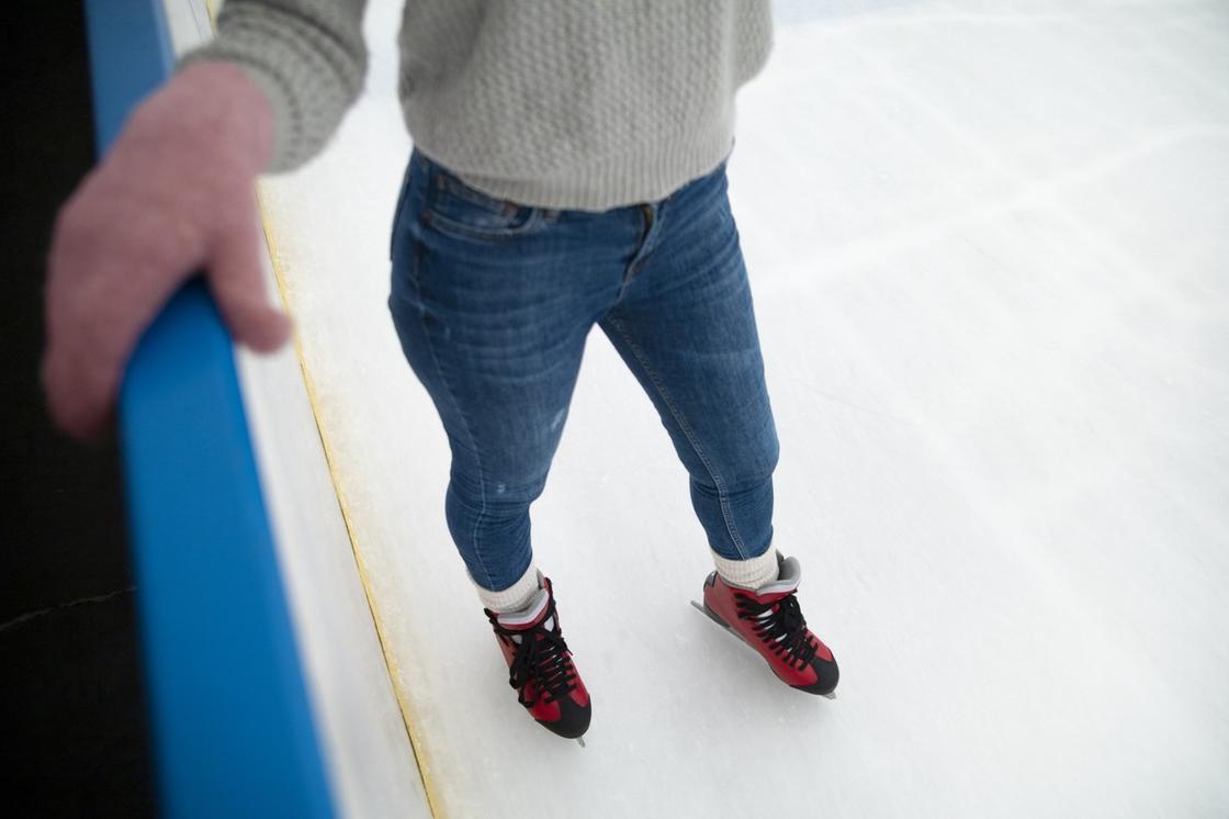 Женщина на коньках стоит и держится за бортик. Она одета в джинсы и свитер