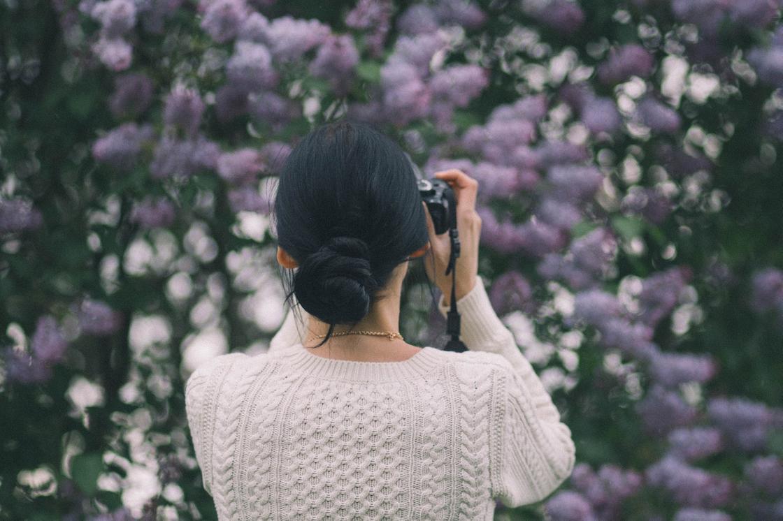 Девушка с низким гладким пучком фотографирует цветы