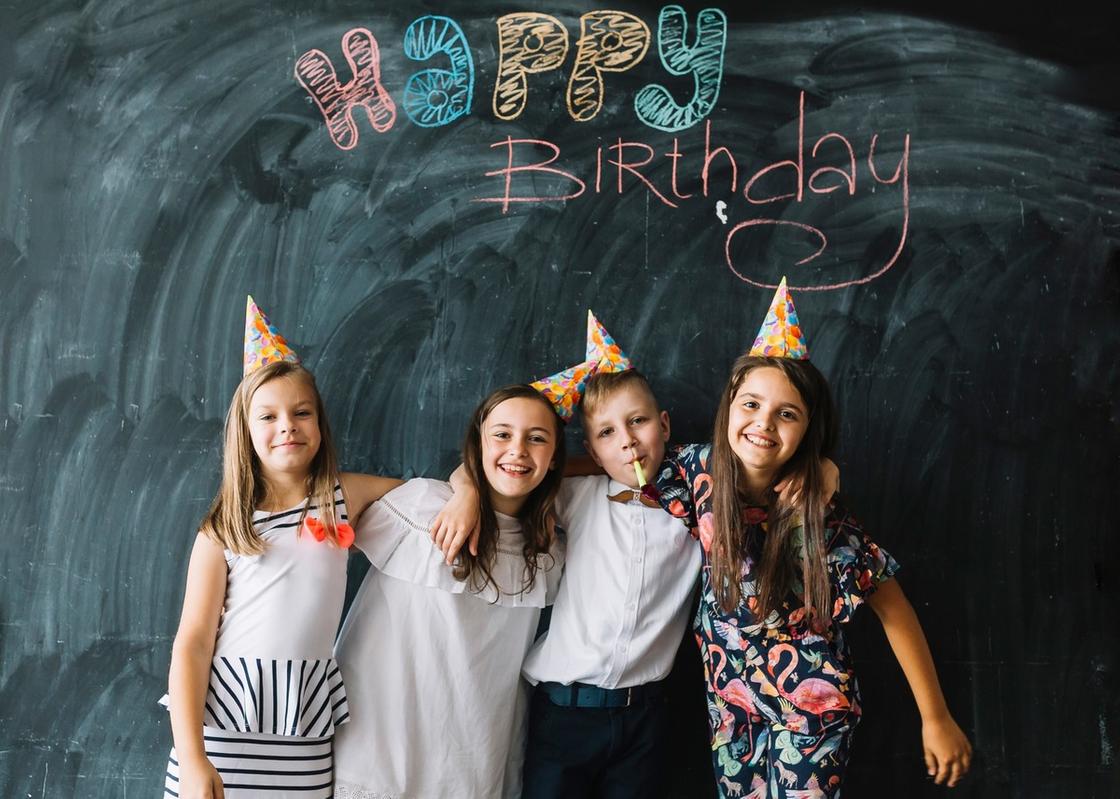 Три девочки и мальчик в колпачках стоят возле школьной доски с надписью «Happy Birthday». Дети обнимаются и улыбаются