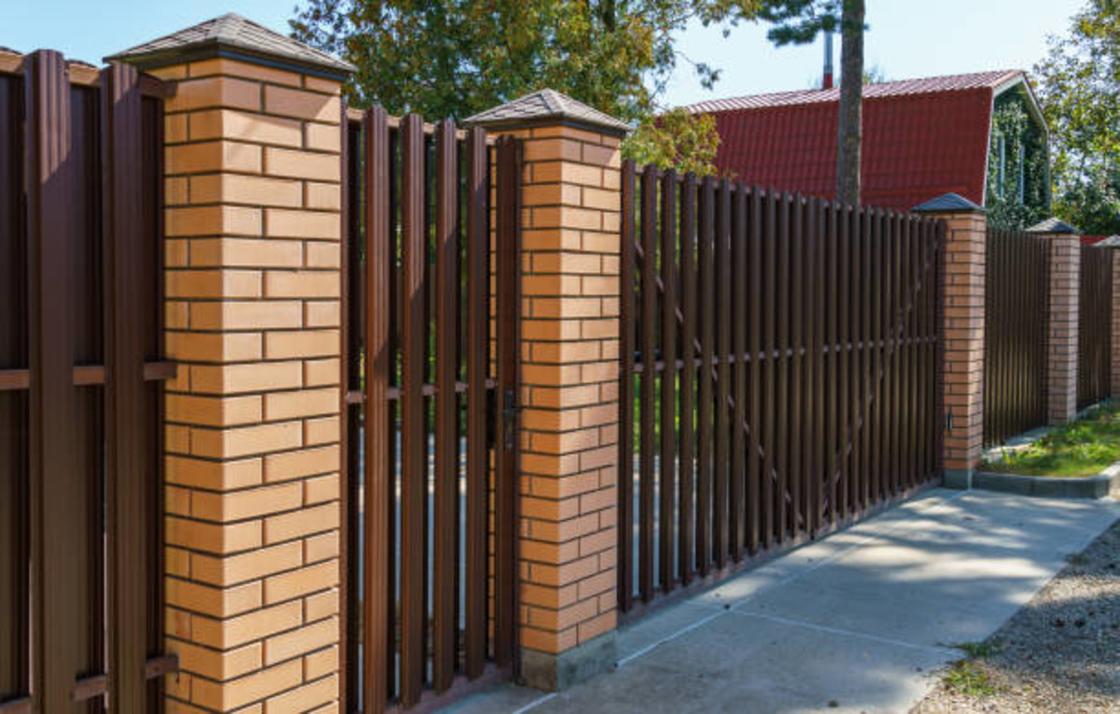 Забор из коричневого металлического штакетника и столбы из кирпича