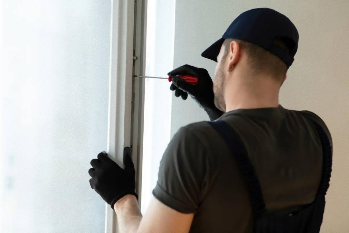 Мужчина в кепке и перчатках проверяет фурнитуру на окне отверткой