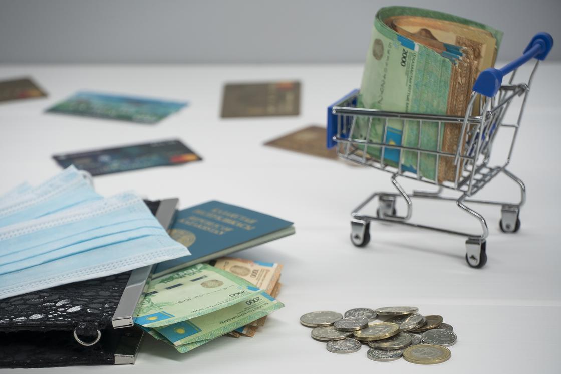 Пачка тенге лежит в миниатюрной тележке между дукоментами, банковскими картами и масками