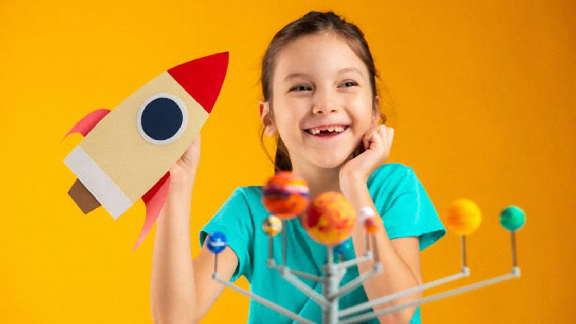 Улыбающаяся девочка в зеленой футболке держит в руках бумажную ракету с красным носом и крыльями. Перед ней стоит сувенир с планетами Солнечной системы