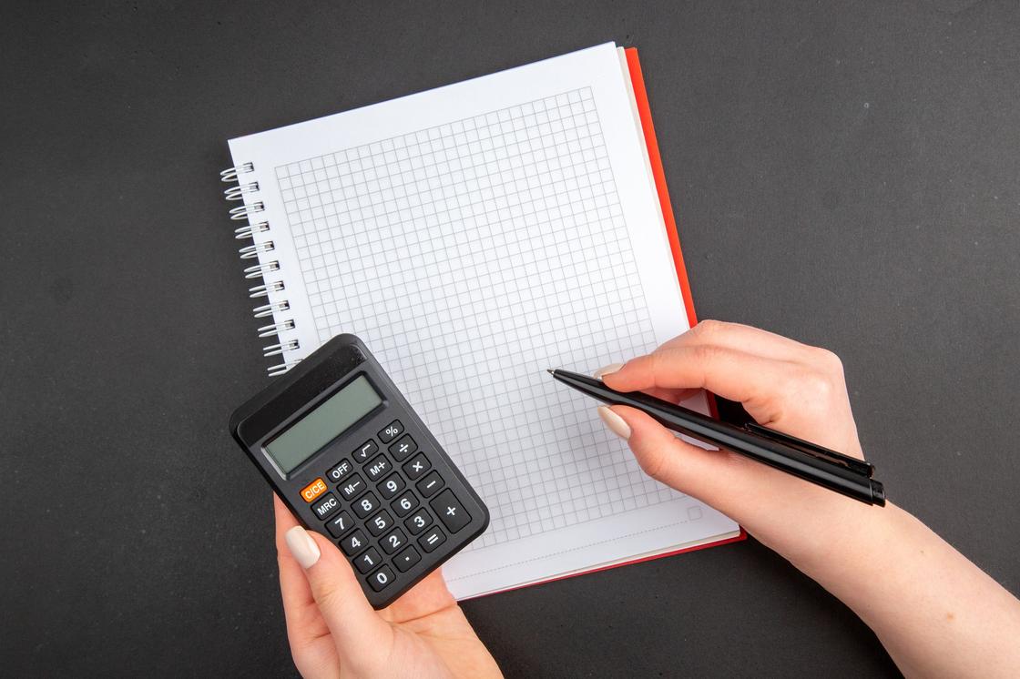 Женская рука держит в право руке ручку над блокнотом, в левой руке калькулятор