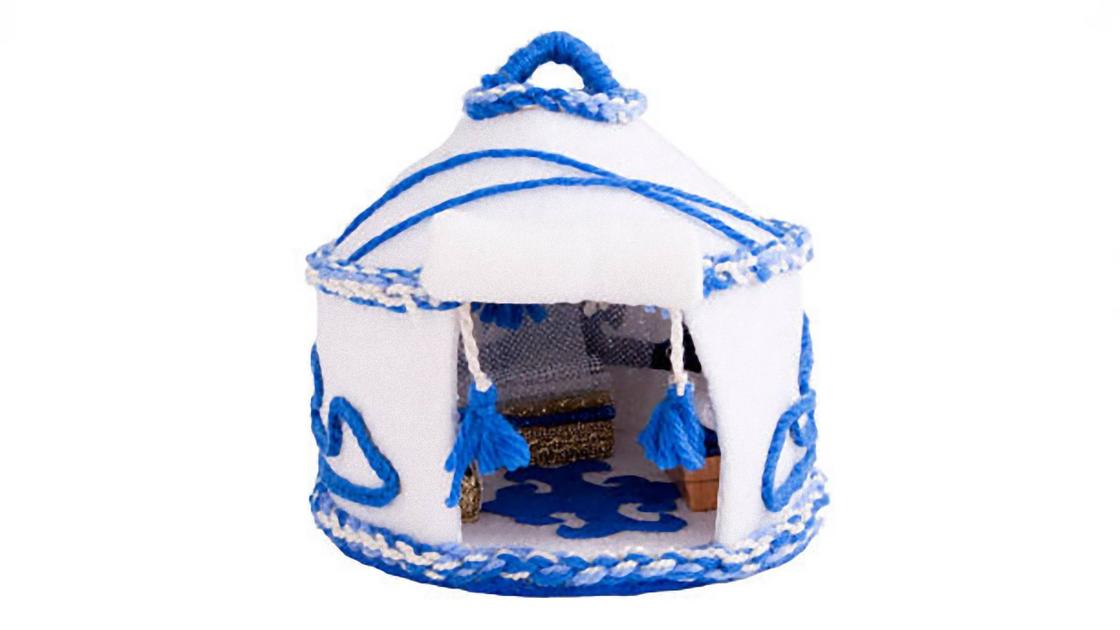 Игрушечная юрта из белого фетра, украшенная синими нитками и косичками. Внутри юрты лежит коврик с орнаментом и стоит сундук