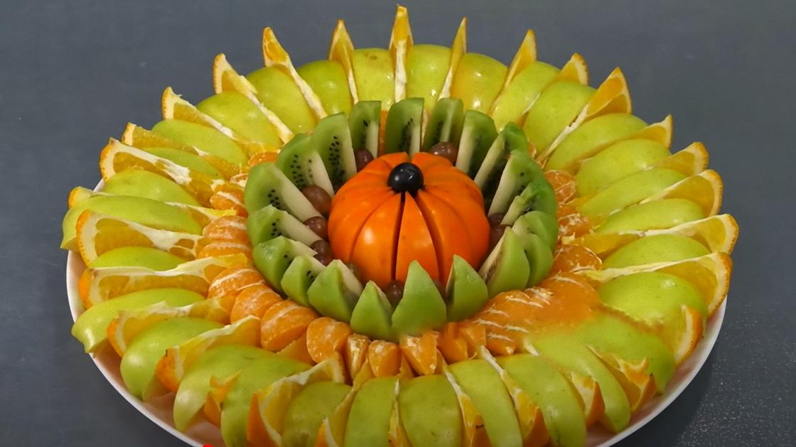 На большом блюде плотно уложены кругами нарезанные зеленые яблоки, апельсины, дольки мандаринов, кусочки киви и разрезанная хурма по центру