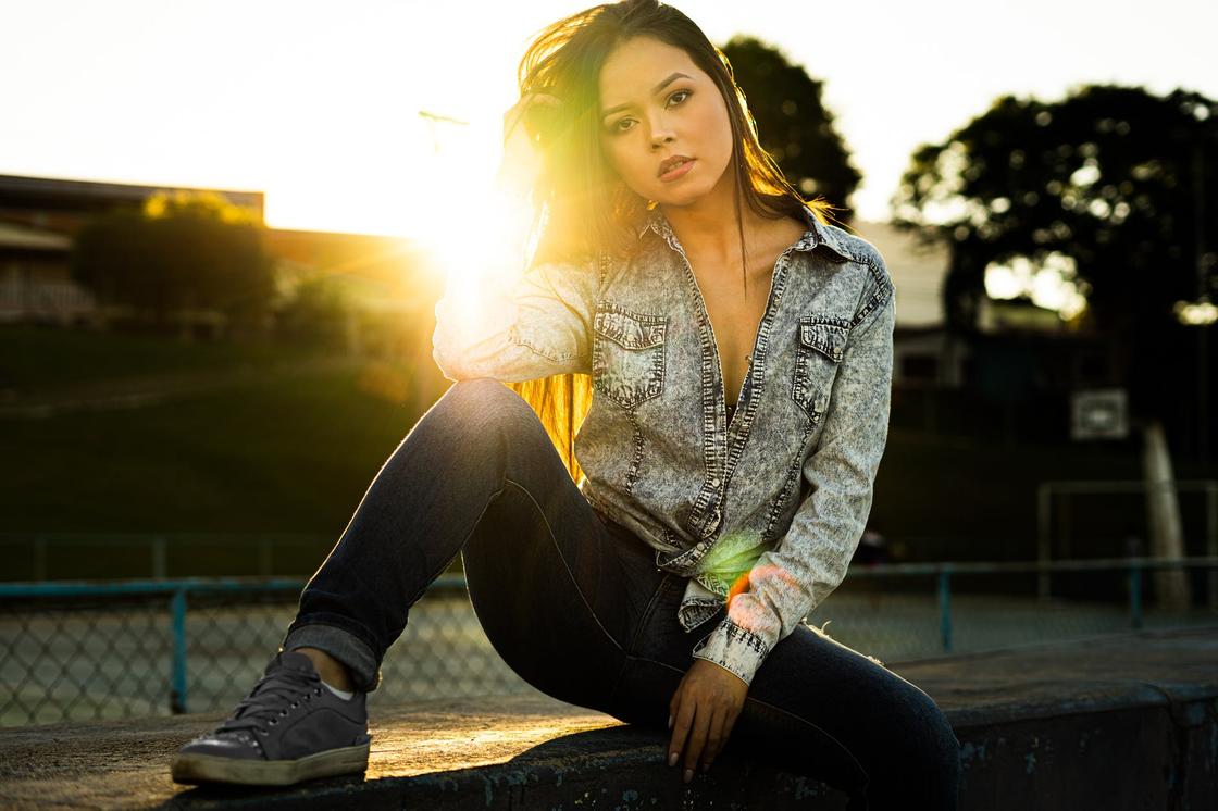 Девушка в джинсовой куртке, джинсах и кедах сидит на фоне закатного солнца