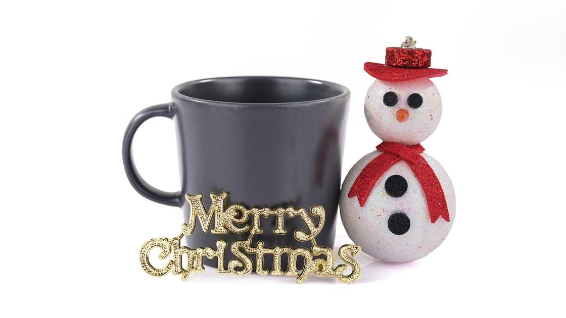 Снеговичок в красной шляпе и шарфике установлен рядом с черной чашкой. Впереди стоит надпись «Merry Christmas»