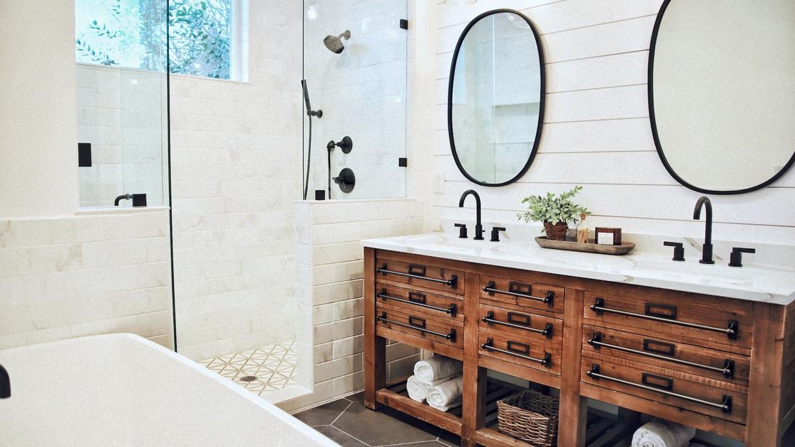 Деревянная тумба с многочисленными ящиками установлена напротив белой ванны, в углу сделана душевая кабина со стеклянной дверью. На тумбе сделаны две раковины с черной фурнитурой, на стене висят овальные зеркала