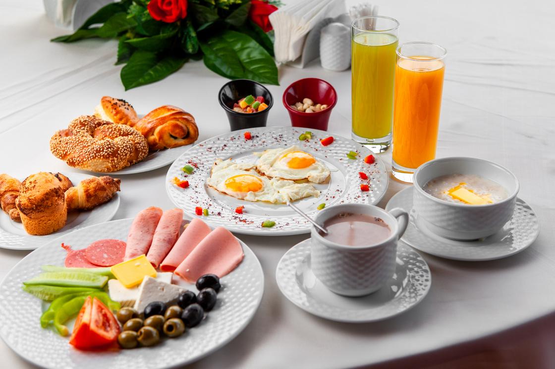 На сервировочном столе жареные яйца, тарелки с выпечкой, соки, кофе, тарелка с холодными закусками