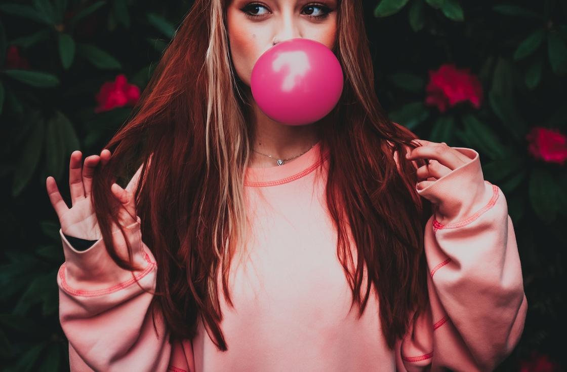 Девушка в розовой одежде надувает пузырь из жвачки