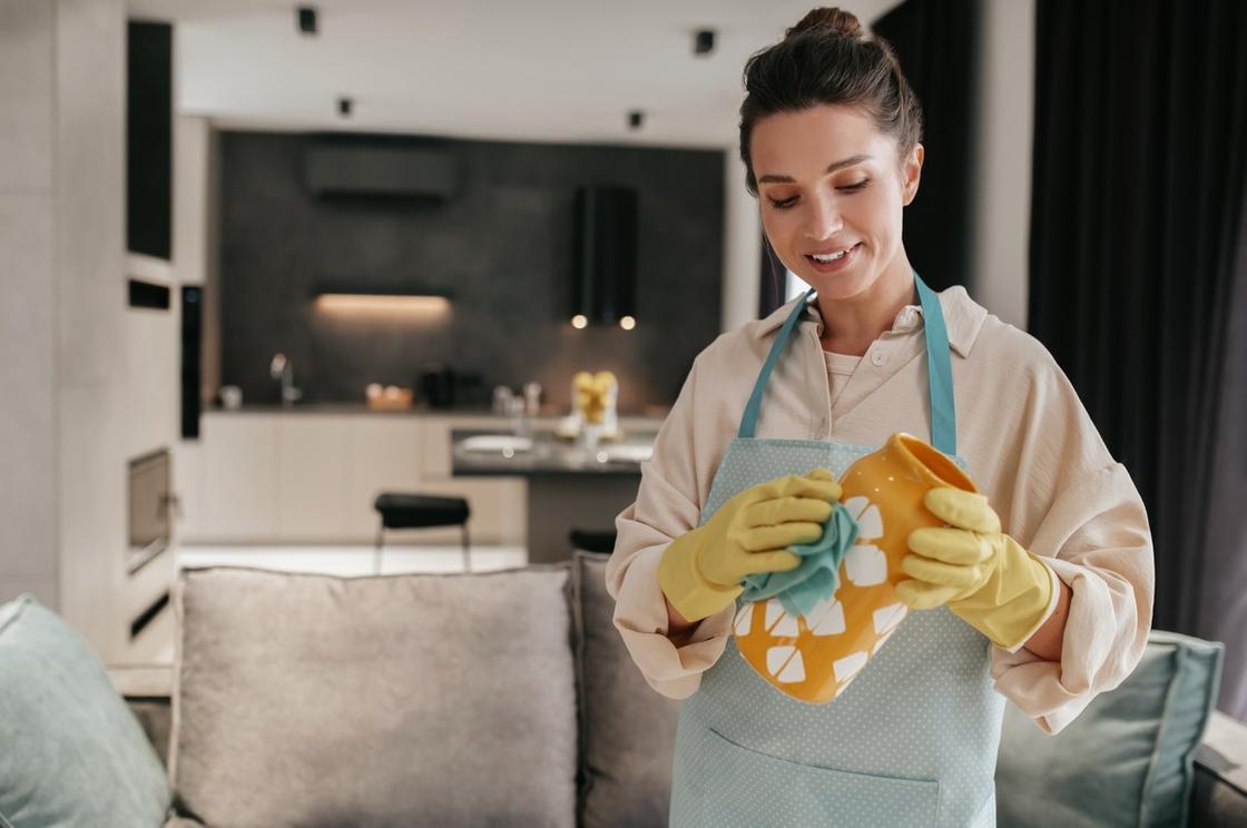 Женщина в фартуке и перчатках вытирает пыль с вазы. Рядом с ней находится мягкий диван