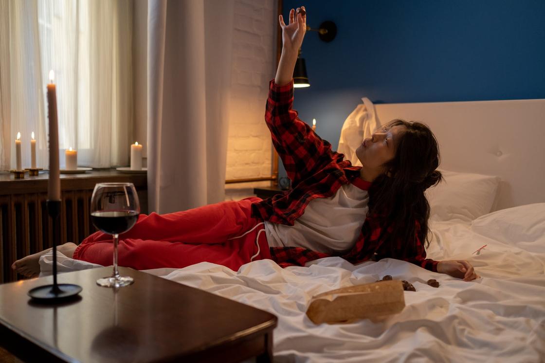 Одинокая женщина в клетчатой рубашке и спортивных штанах лежит в кровати рядом с бокалом вина