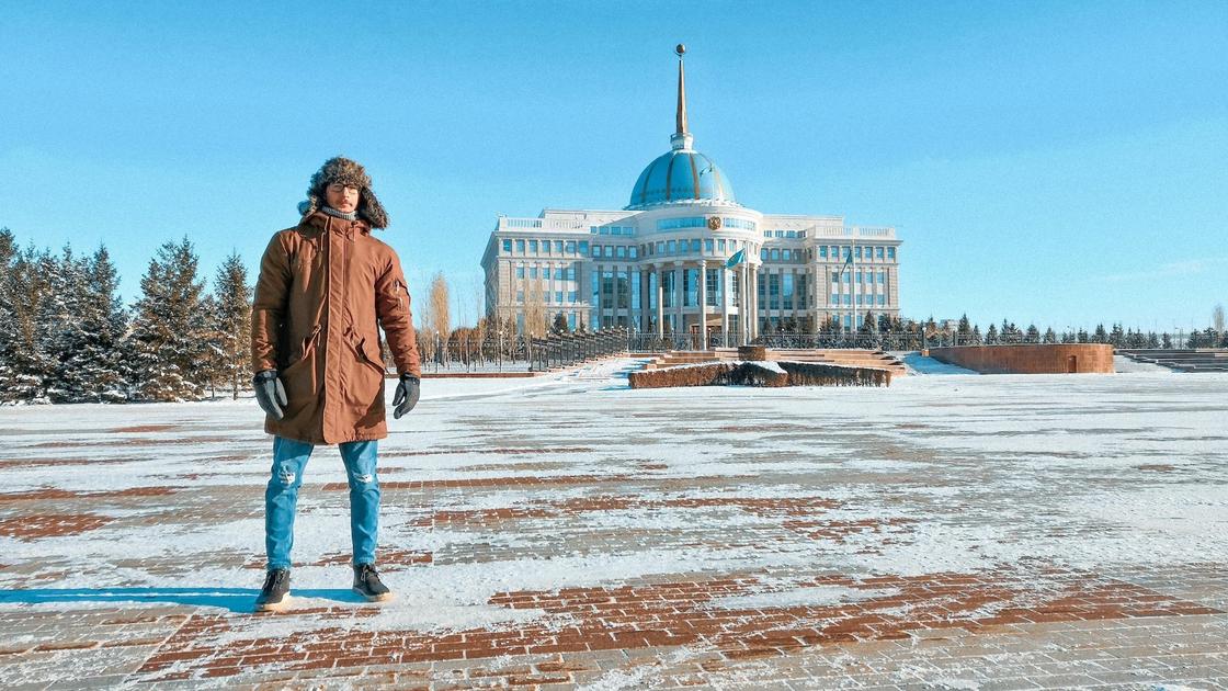 Мужчина в теплой зимней куртке, перчатках и шапке-ушанке стоит на большой площади возле красивого дворца. Под ногами у него плитка, слегка покрытая снегом
